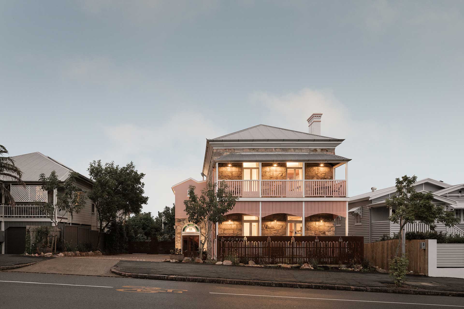 A large two-storey house painted pink with white trim, featuring balconies on both levels. Surrounding trees and shrubs provide greenery, and a wooden fence encloses the front yard. MIss midgleys is positioned on james st in new farm, brisbane