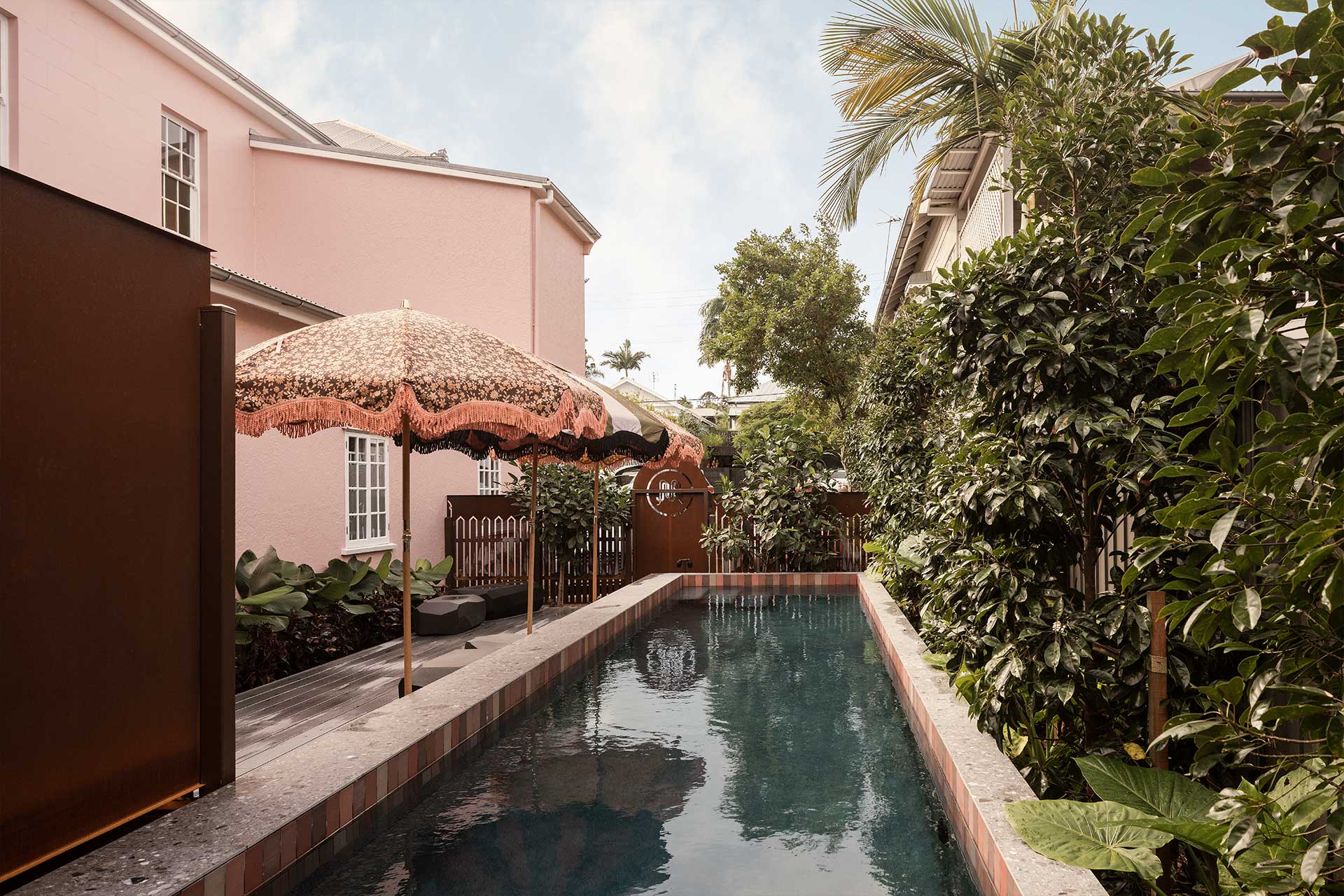 A tranquil garden setting featuring a narrow pool bordered by lush greenery and palm trees. A decorative umbrella provides shade beside the water, with soft pink walls of a house visible in the background. This scene reflects the aesthetic of Miss Midgley's New Farm.