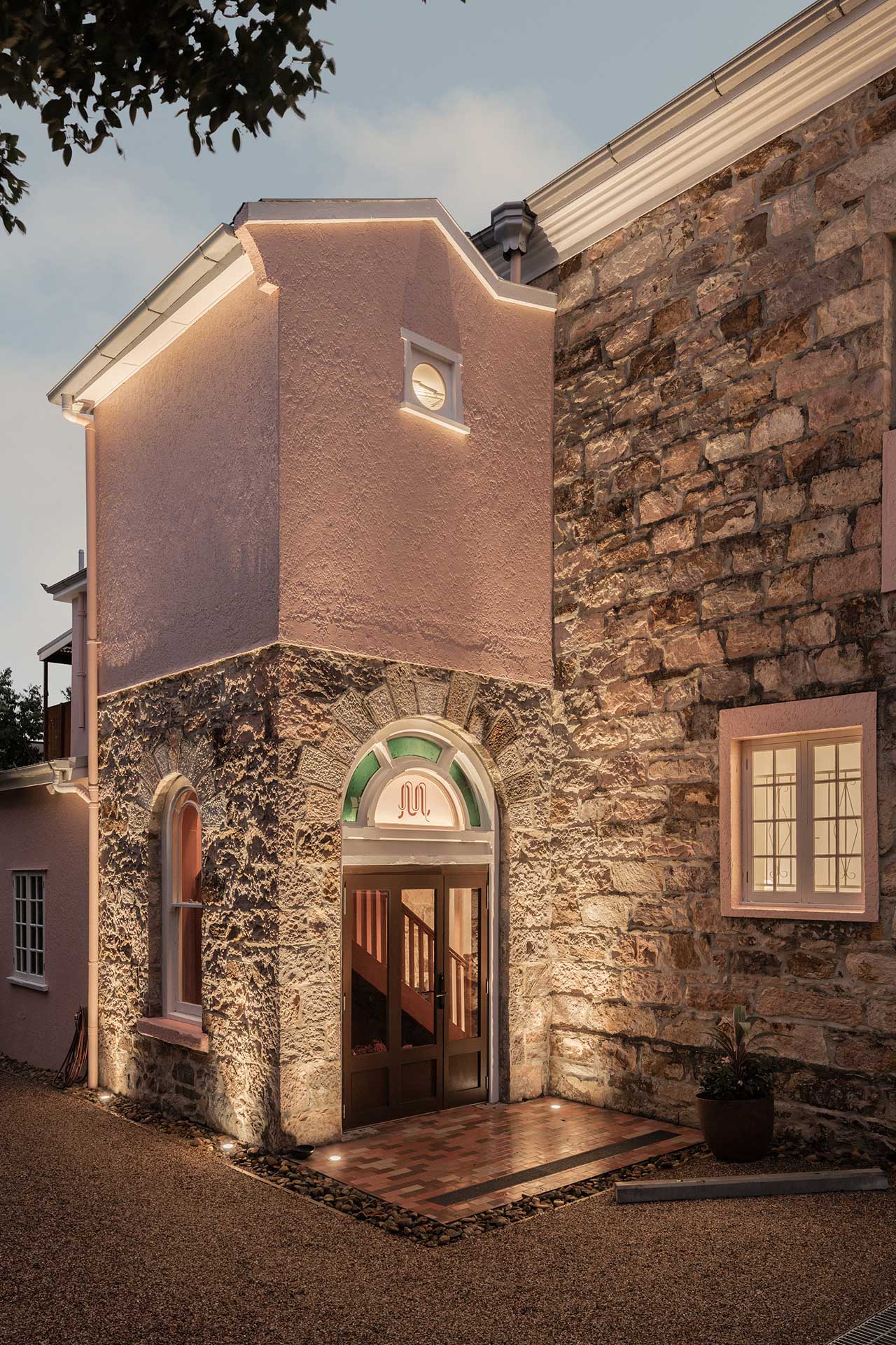 The entrance of a building with a mix of pink plaster and stone walls, featuring an arched doorway framed with green and white accents. Soft lighting highlights the architecture, and a gravel path leads towards the entrance.